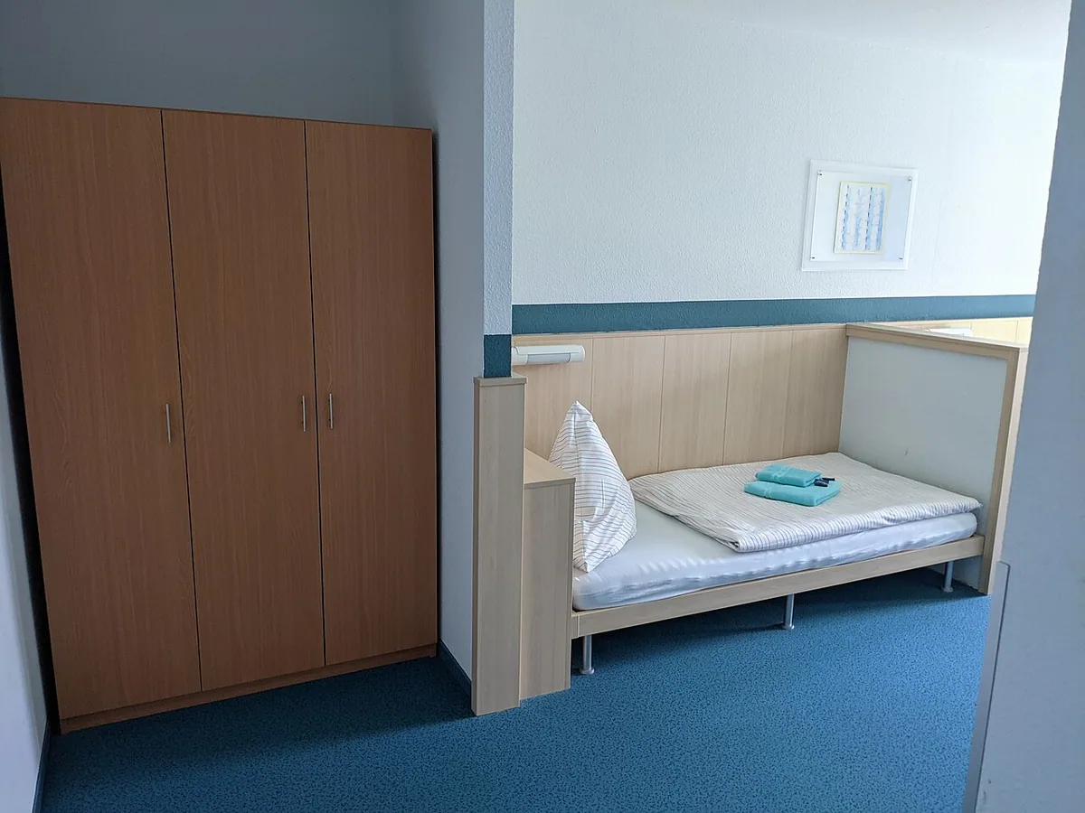 A single bed with folded towels beside a wooden wardrobe in a simple guest room
