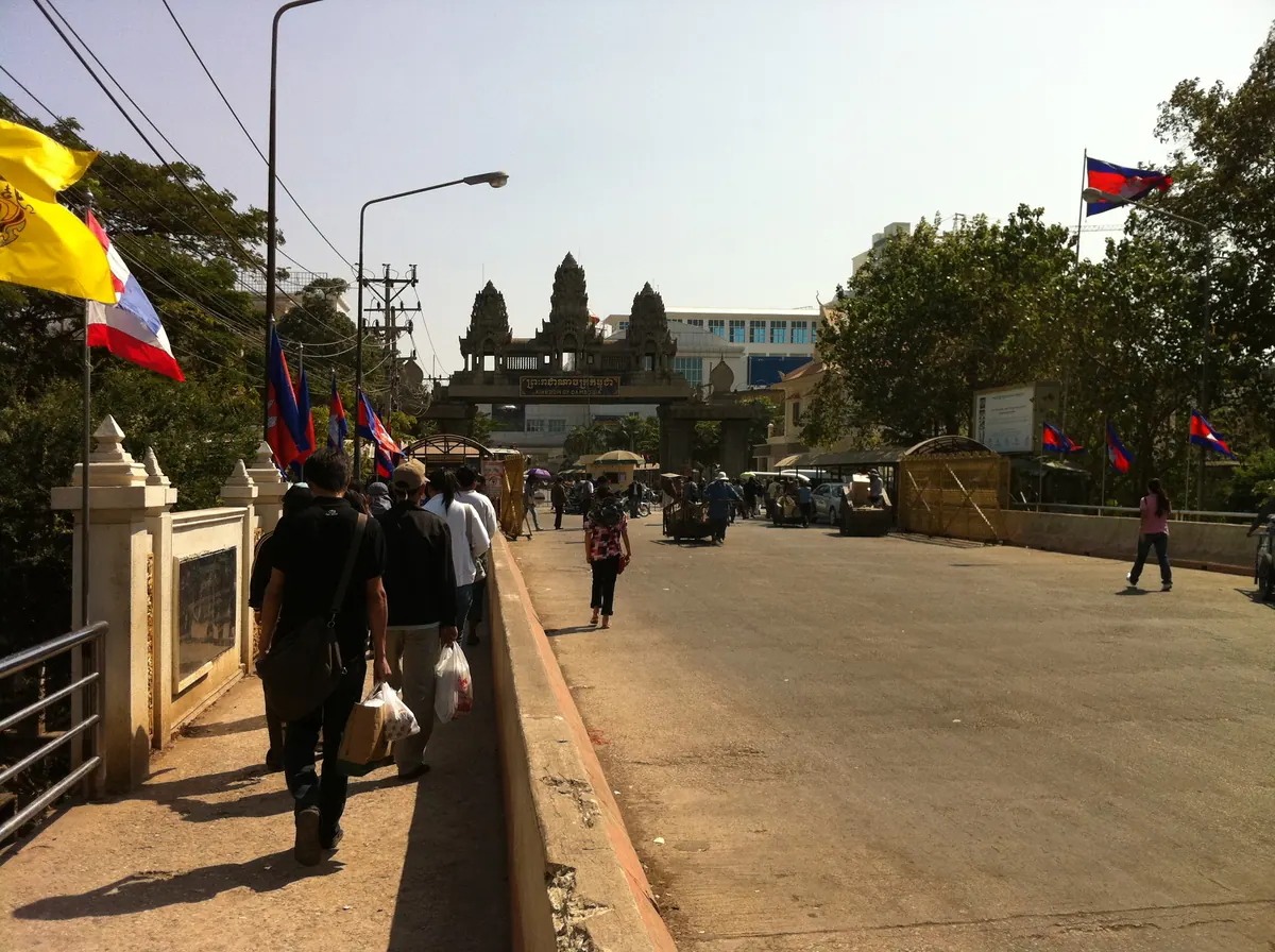 Travellers walking across a land border crossing between Thailand and Cambodia, with both flags flying