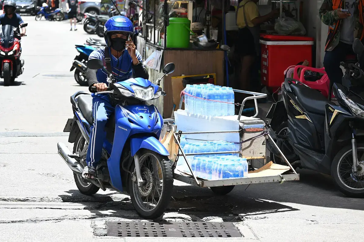 A rider in a blue helmet on a blue Honda scooter, towing a trailer loaded with packs of bottled water along a street